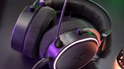 Na ciemnym biurku leżą czarne słuchawki z mikrofonem, klawiatura oraz perforowana mysz z podświetleniem RGB.