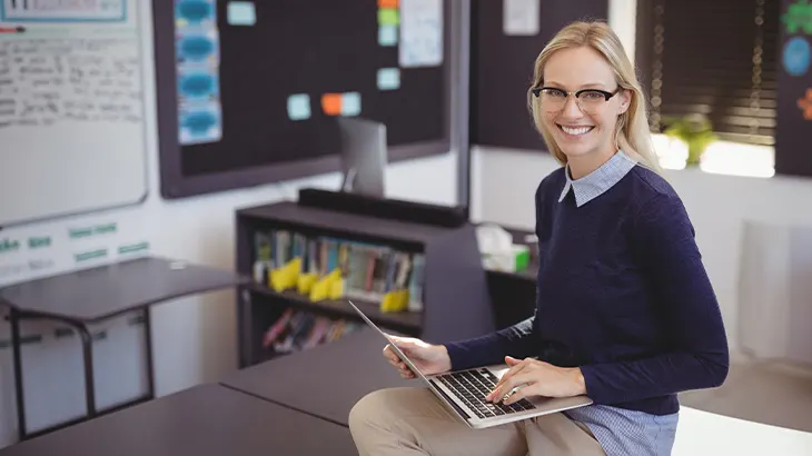 Kobieta w okularach siedzi na biurku w klasie i używa laptopa. W tle tablica, regał z książkami i inne biurka.