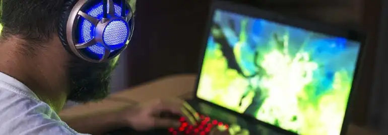 Osoba w słuchawkach z niebieskim światłem gra na laptopie z podświetlaną na czerwono klawiaturą. Ekran laptopa wyświetla grafikę w kolorach zielonym i niebieskim.