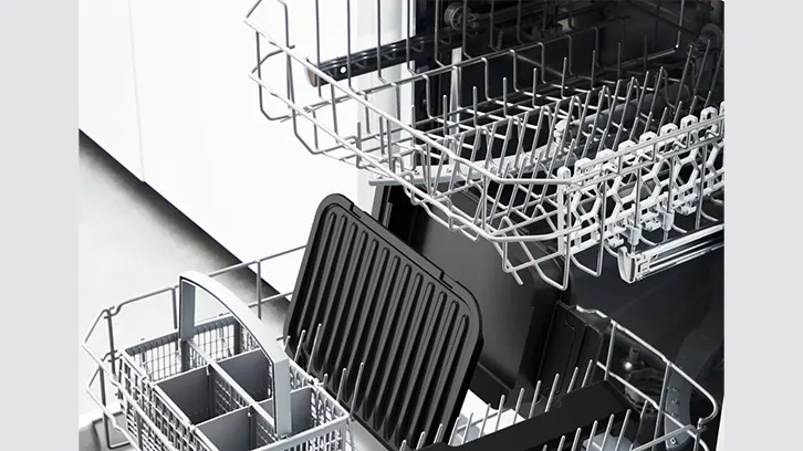 Wnętrze zmywarki z metalowymi koszami na naczynia, w dolnym koszu czarna blacha do pieczenia i pojemnik na sztućce.