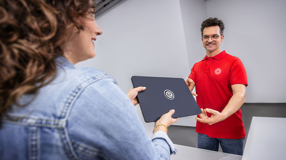 Kobieta w kurtce dżinsowej odbiera czarny laptop od mężczyzny w czerwonej koszulce polo z logo.