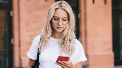 Kobieta w okularach czyta na czerwonym telefonie. Nosi białą koszulkę. W tle budynek z cegły z czarnymi drzwiami.