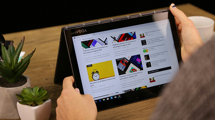 Osoba używa tabletu Yoga na stole, obok doniczek z roślinami. Ekran tabletu wyświetla stronę internetową. W tle widać biały kubek.