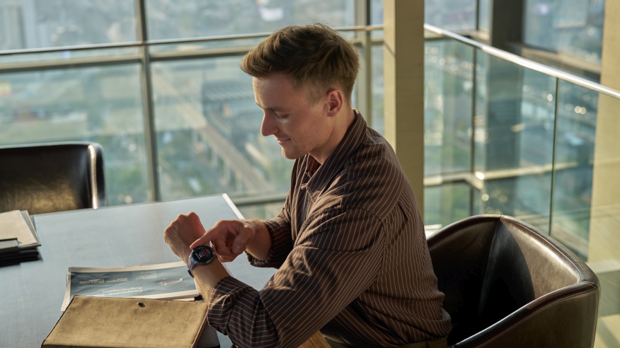Mężczyzna w koszuli w paski sprawdza zegarek na tle okna z widokiem na miasto. Na stole leżą dokumenty.