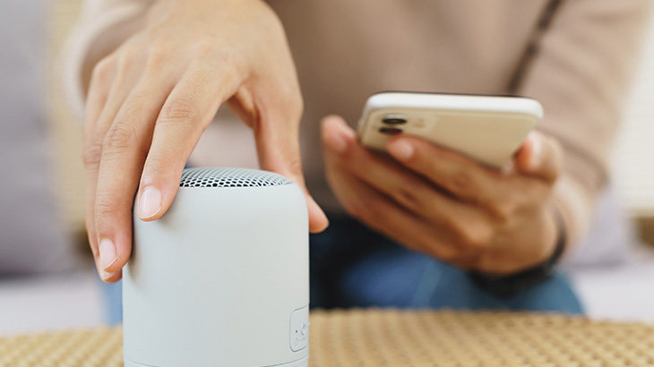 Osoba trzyma biały głośnik i telefon komórkowy na plecionym stoliku.