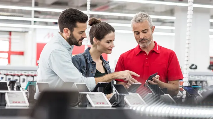 W sklepie, mężczyzna i kobieta oglądają telefony z sprzedawcą w czerwonej koszuli, który pokazuje im produkt.