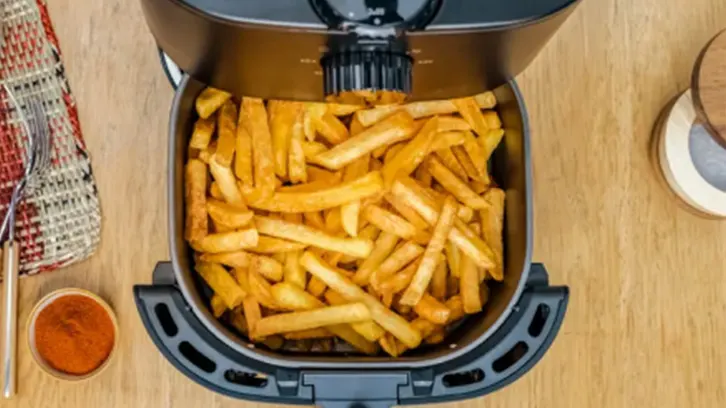 Frytkownica wypełniona frytkami na drewnianym stole, obok sos, serwetka i widelec.