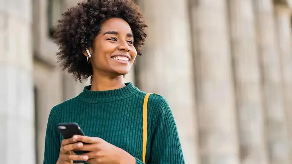 Kobieta w zielonym swetrze trzyma telefon. Ma słuchawki, a przez ramię przewieszony jest żółty pasek torby. W tle widać kolumny.