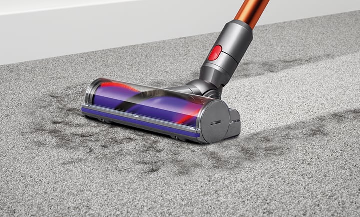 Odkurzacz na szarej wykładzinie, czyszczący zabrudzenia. Rura odkurzacza jest pomarańczowo-szara, a szczotka przezroczysta z fioletowym akcentem.