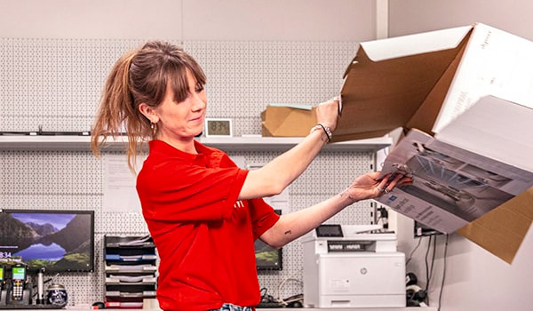 Kobieta w czerwonej koszulce wyjmuje plakat z kartonowego pudła w biurze z drukarką, półkami i monitorem.