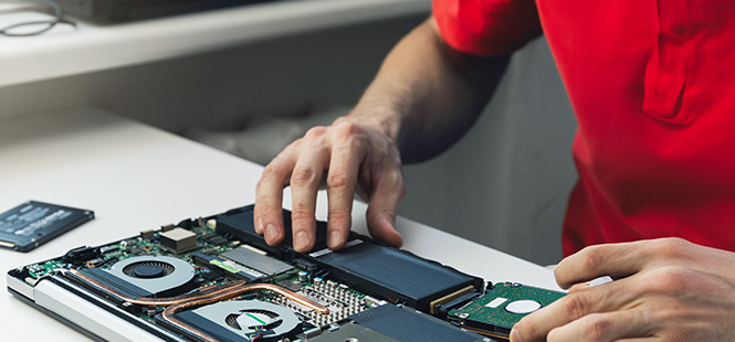 Osoba w czerwonej koszulce naprawia laptopa. Na białym stole rozłożony jest laptop, widoczne są elementy wewnętrzne.