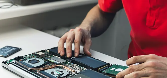 Osoba w czerwonej koszulce naprawia laptopa. Na białym stole rozłożony jest laptop, widoczne są elementy wewnętrzne.