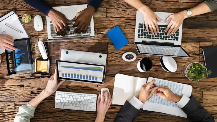 Na drewnianym stole leżą laptopy, tablety, notatniki, myszki komputerowe, klawiatura i roślina w doniczce. Wokół stołu widać ręce osób pracujących.