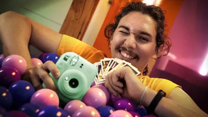 Uśmiechnięty mężczyzna w żółtej koszulce leży w basenie z kulkami, trzyma zielony aparat i zdjęcia.