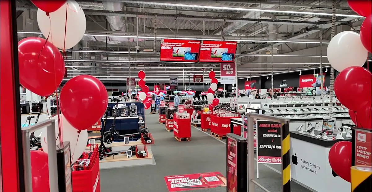 Wnętrze sklepu z elektroniką, dekoracje z czerwonych i białych balonów. Widoczne regały z towarami i reklamy.