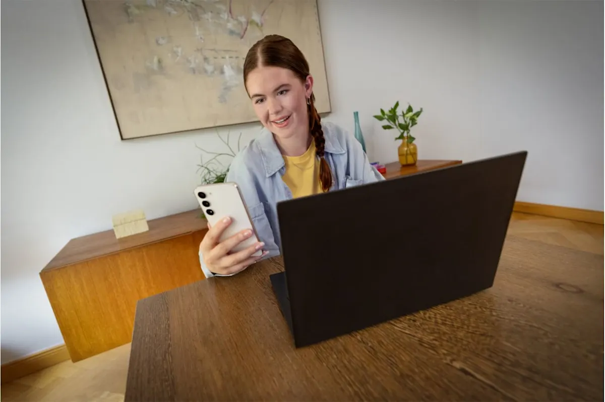 Kobieta siedzi za drewnianym biurkiem z laptopem i trzyma w ręku smartfon. Za nią komoda i obraz na białej ścianie.