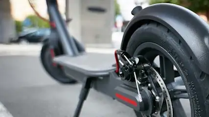 Widok na czarno-szarą hulajnogę elektryczną na ulicy, z widocznym tylnym kołem, błotnikiem, światłem odblaskowym i hamulcem tarczowym.