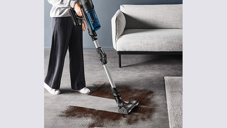 Osoba odkurza brudną podłogę odkurzaczem. W tle szara kanapa i dywan.