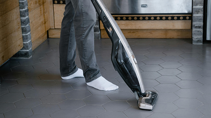 Osoba w szarych spodniach odkurza szarą podłogę odkurzaczem pionowym, na tle drewnianych szafek i metalowych urządzeń.