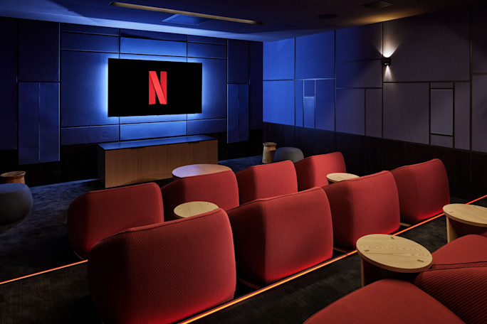 Kameralna sala kinowa z ciemnoniebieskimi ścianami i rzędami wygodnych, czerwonych foteli, na której ekranie wyświetlane jest logo Netflix.