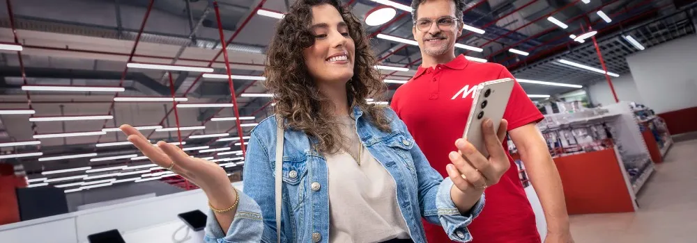 Kobieta w dżinsowej kurtce i mężczyzna w czerwonej koszulce oglądają telefon w sklepie. W tle oświetlenie i półki z produktami.