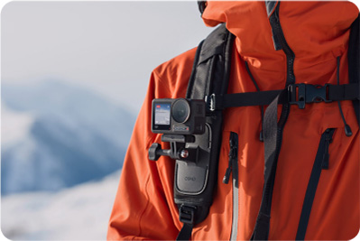 Osoba w pomarańczowej kurtce z kamerą przymocowaną do paska plecaka na tle gór.