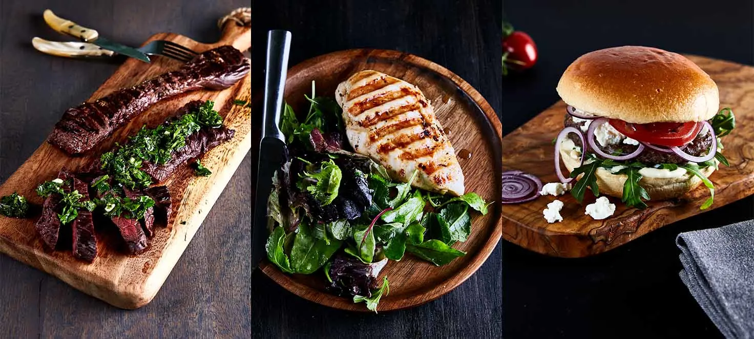 Zdjęcie przedstawia trzy potrawy: stek z ziołami, kurczaka z sałatą i burgera z warzywami, podane na drewnianych deskach.
