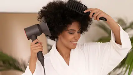 Kobieta w szlafroku suszy włosy suszarką i czesze je czarną szczotką na tle jasnego pomieszczenia i rośliny.