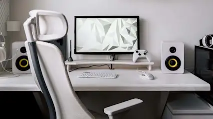Białe biurko z komputerem, głośnikami, słuchawkami, padem i roślinami. Krzesło biurowe stoi przed biurkiem.