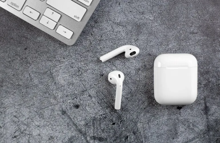 Dwa białe słuchawki bezprzewodowe i etui obok klawiatury na szarym tle.