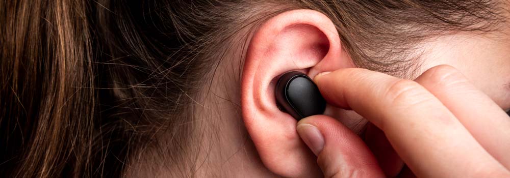 Czarny, bezprzewodowy słuchawka w uchu osoby, trzymana palcem. Widoczny fragment ucha i włosów.