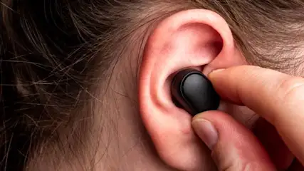 Czarny, bezprzewodowy słuchawka w uchu osoby, trzymana palcem. Widoczny fragment ucha i włosów.