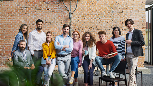 Grupa ludzi stoi na tle ceglanej ściany. Niektórzy siedzą na krzesłach lub opierają się o ścianę. Ubrani są w różnorodne stroje.