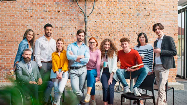 Grupa ludzi stoi na tle ceglanej ściany. Niektórzy siedzą na krzesłach lub opierają się o ścianę. Ubrani są w różnorodne stroje.