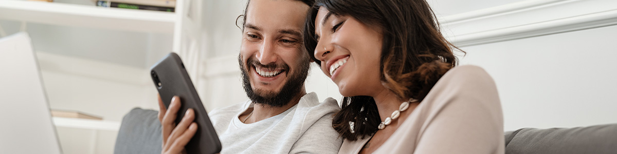 Uśmiechnięta para siedzi na szarej sofie i patrzy na czarny telefon komórkowy trzymany przez mężczyznę w białej koszulce.