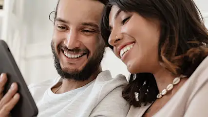 Uśmiechnięta para siedzi na szarej sofie i patrzy na czarny telefon komórkowy trzymany przez mężczyznę w białej koszulce.