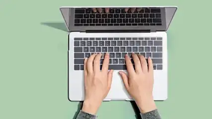 Widok z góry na laptopa na zielonym tle. Dłonie piszą na klawiaturze. Ekran laptopa jest częściowo otwarty.