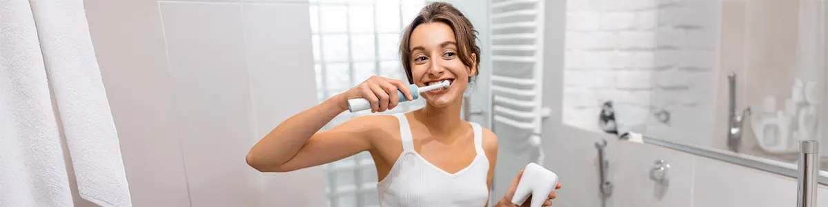Kobieta w białym topie myje w łazience zęby białą szczoteczką elektryczną z główką rotacyjną. W drugim ręku trzyma kubek w kształcie zęba trzonowego.
