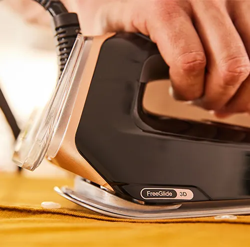 Czarno-złote żelazko na żółtej tkaninie, prasowane przez osobę. Widoczny kabel i napis "FreeGlide 3D".