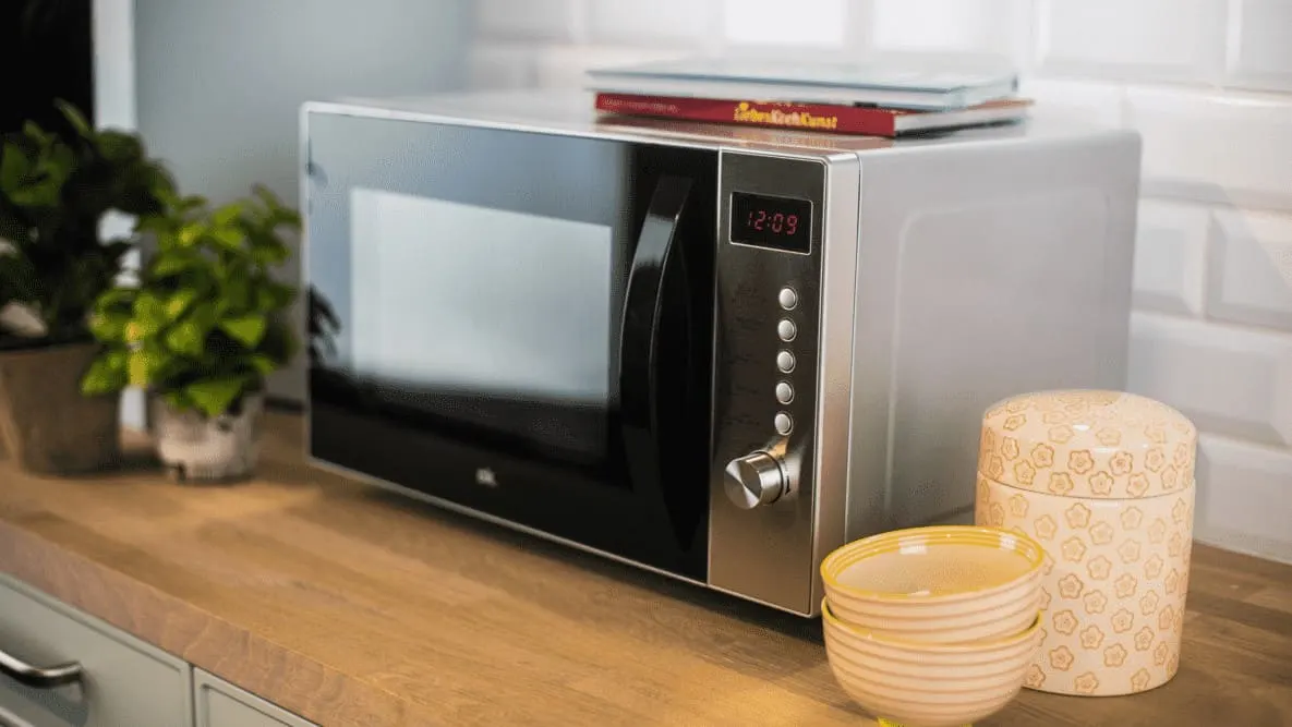 Mikrofalówka na blacie, obok miski i pojemnik. Na mikrofalówce leżą książki. W tle rośliny doniczkowe i białe kafelki.