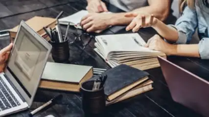 Na ciemnym stole leżą laptopy, notatniki, książki i przybory do pisania. Wokół stołu siedzą osoby, pracują lub dyskutują.