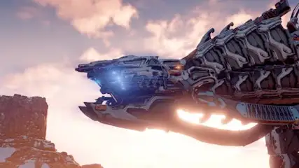 Na zdjęciu widać mechanicznego stwora na tle zachodzącego słońca i skalistego krajobrazu. Góry i drzewa są częściowo pokryte śniegiem.