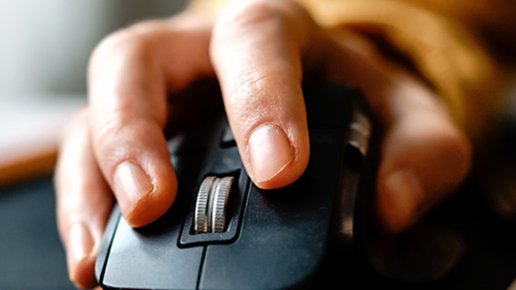 Dłoń naciska na czarny przycisk myszy komputerowej z metalową rolką przewijania na rozmytym tle.