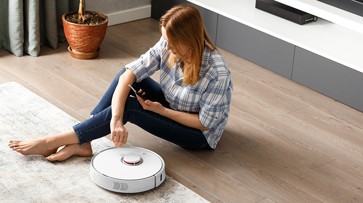 Kobieta siedzi na podłodze obok robota sprzątającego, trzyma telefon. W tle podłoga, dywan, szafka RTV i roślina w doniczce.