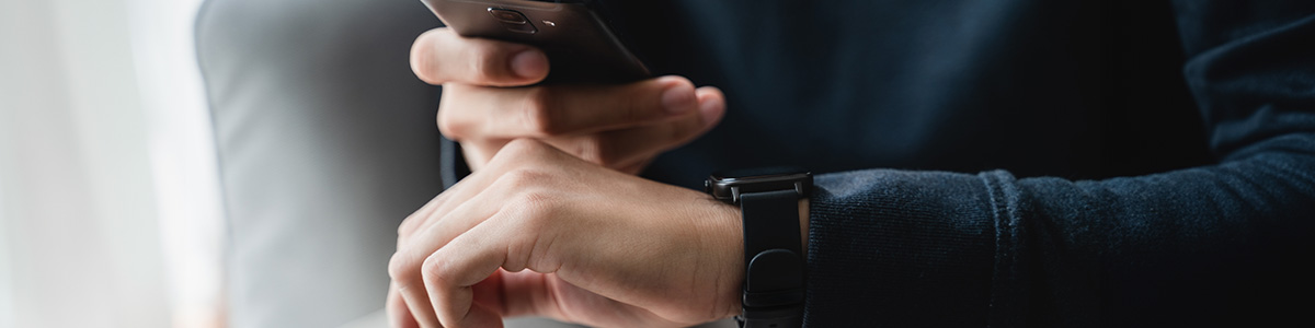 Osoba trzyma czarny telefon i ma na nadgarstku czarny zegarek na tle szarego krzesła i białej ściany.