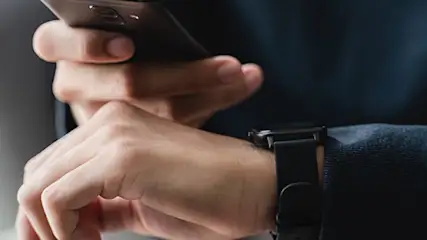 Osoba trzyma czarny telefon i ma na nadgarstku czarny zegarek na tle szarego krzesła i białej ściany.