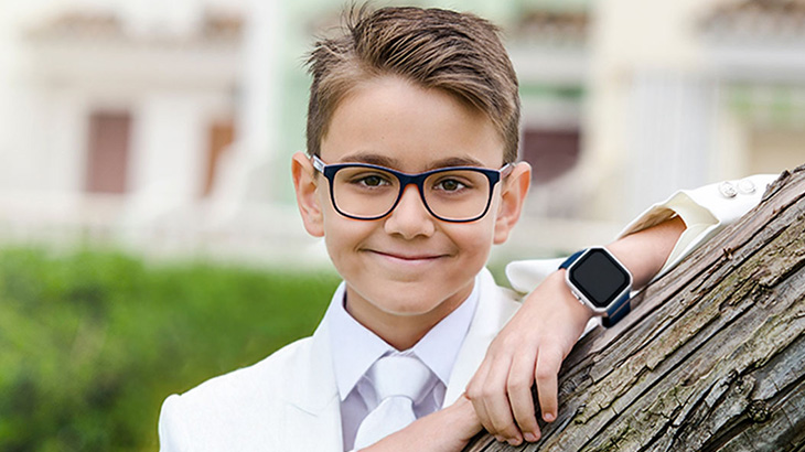 Chłopiec w okularach, ubrany w biały garnitur i krawat, opiera się o brązowe drzewo. Na nadgarstku ma niebieski zegarek.