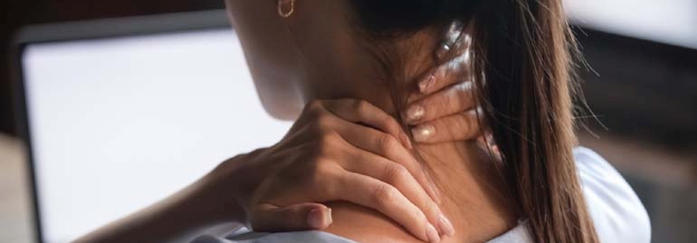 Kobieta w białej koszuli masuje szyję dłońmi na tle ekranu komputera i ciemnego otoczenia.