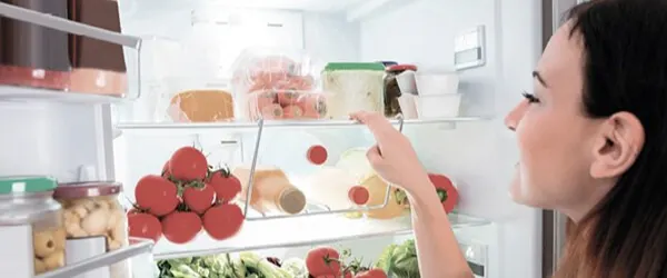 Kobieta sięga do otwartej lodówki, w której są pomidory, sałata, sery i inne produkty spożywcze w pojemnikach i słoikach.