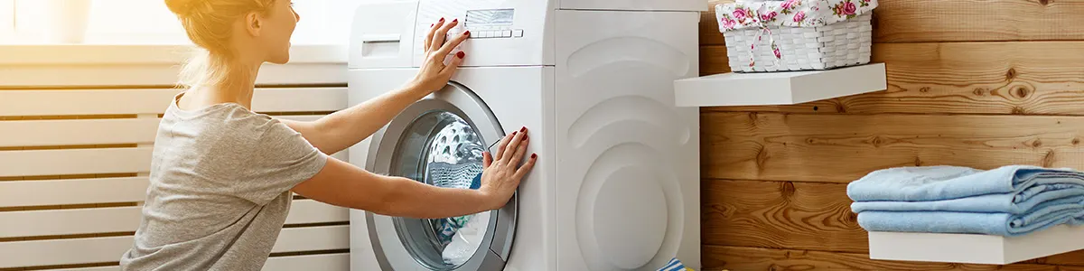 Kobieta włącza białą wąską pralkę ładowaną od przodu. Z boku czyste tkaniny.